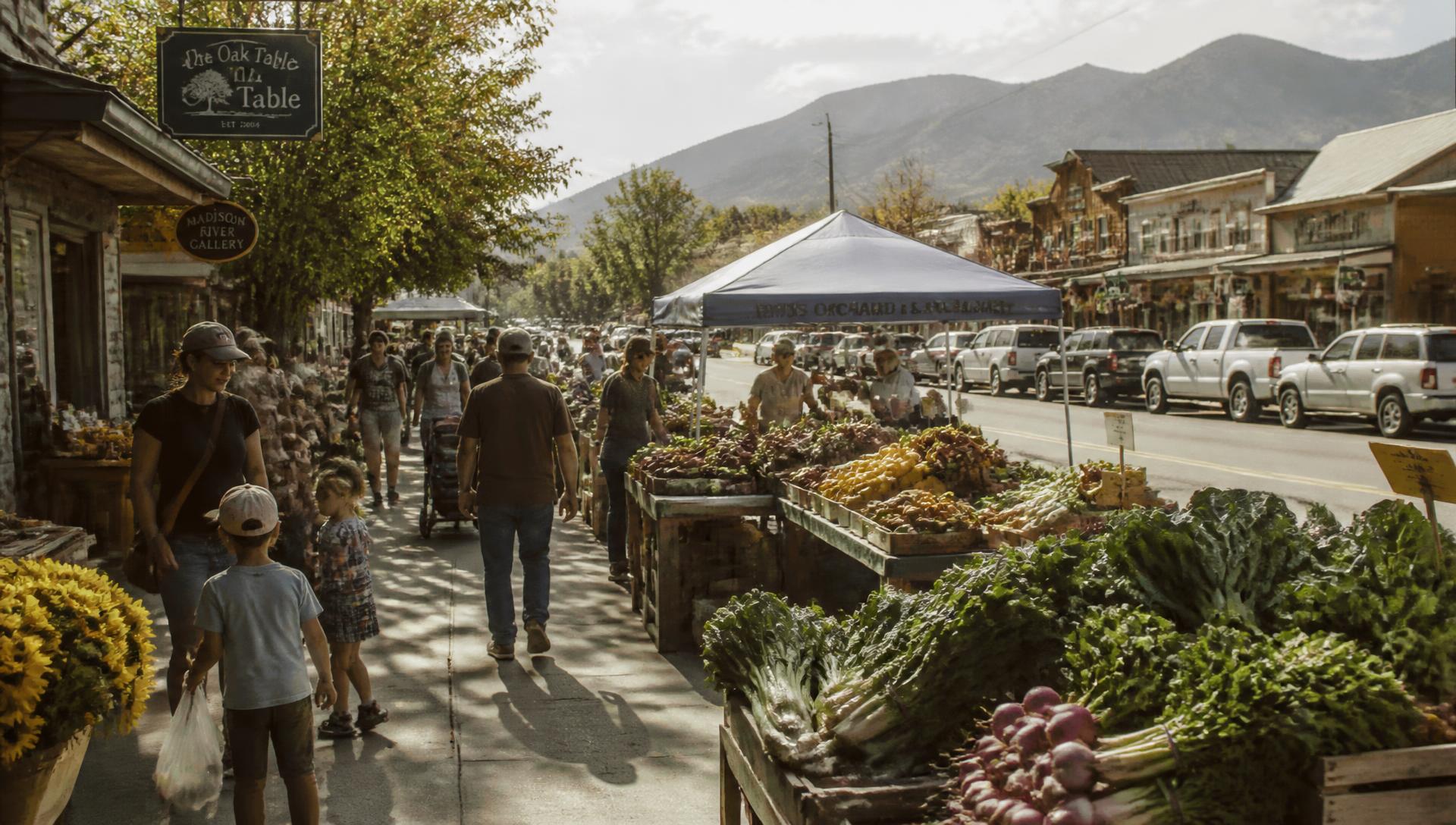 Families walking a small town main street market with farm produce and mountains beyond